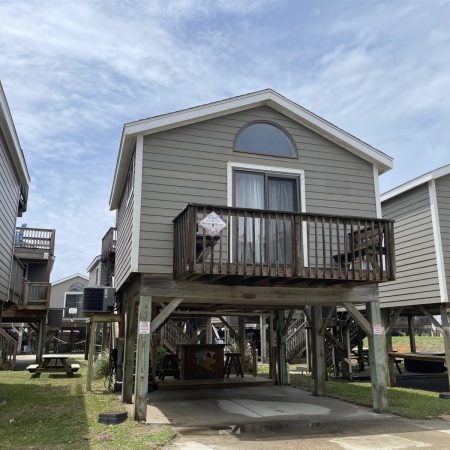 Gray beach house on stilts with a small balcony and a paved area beneath, surrounded by other similar houses.
