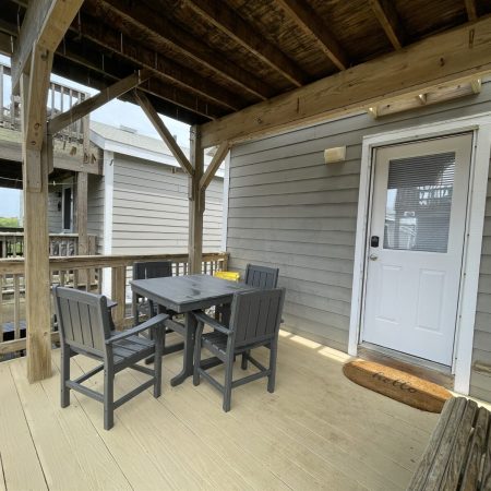 Wooden deck with a table, four chairs, and a doormat in front of a white door on a gray-sided building.