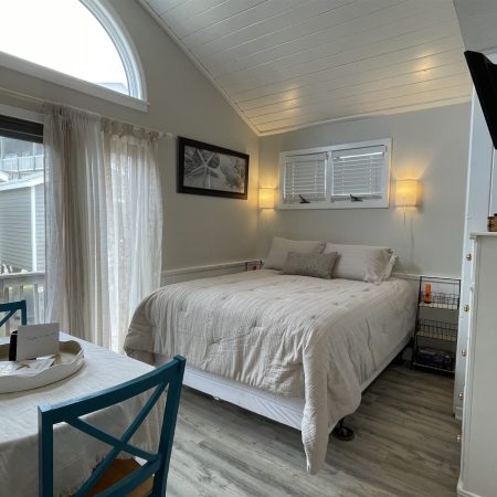 Small, bright bedroom with a double bed, blue chairs at a table, wall TV, and large window with sheer curtains.