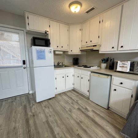 Small kitchen with white cabinets, appliances, wood-patterned floor, and a door with window blinds.