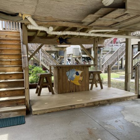 Wooden bar with two stools under stairs in an outdoor area; a painted fish decorates the bar front.