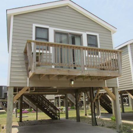 Elevated gray beach house with a small balcony and stairs leading to a parking area underneath.