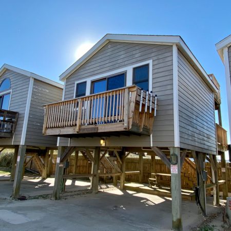 Elevated beach house with wooden balcony and stairs, set on stilts above a paved area under a clear blue sky.