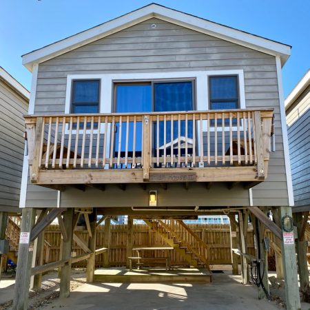 Elevated beach house with a wooden balcony, sliding glass doors, and parking space underneath on a sunny day.