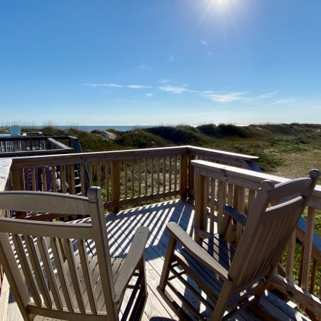 Two wooden rocking chairs on a sunny deck overlook grassy dunes and a clear blue sky by the beach.
