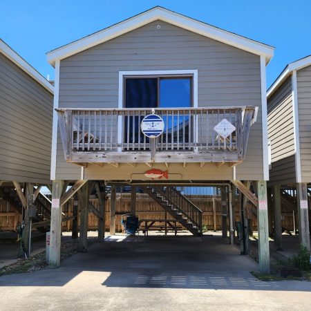 A beige elevated beach house with a wooden balcony and stairs underneath, supported by stilts, on a sunny day.