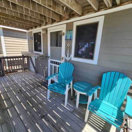 Two turquoise Adirondack chairs and a small table sit on a wooden porch outside a beige house with white trim.
