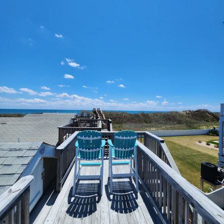 Two blue chairs on a wooden deck overlook grassy dunes and the ocean under a clear blue sky.