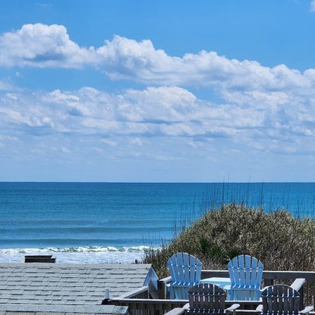Two white Adirondack chairs face the ocean, with grassy dunes and rooftops in the foreground under a blue sky.