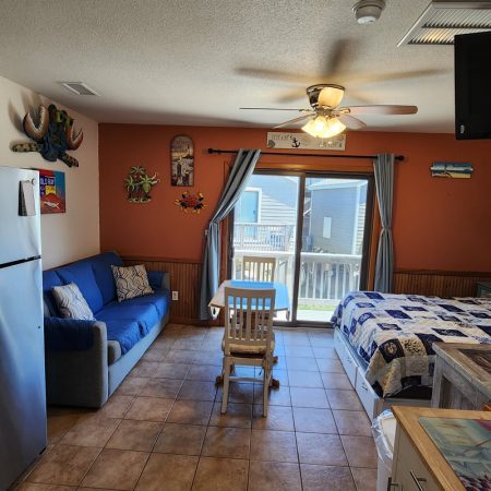 Studio room with a blue sofa, dining table, bed, fridge, wall decor, and window showing a balcony view.