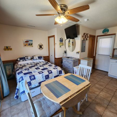 Small, tidy studio room with a bed, table and chairs, dresser, TV on wall, and a ceiling fan above tile flooring.