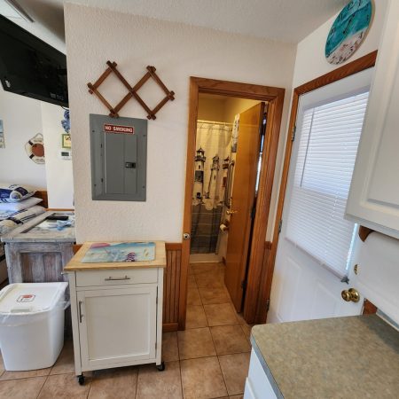 View of a small kitchen area leading to a bathroom with a shower curtain, beige tile floor, and wooden trim.