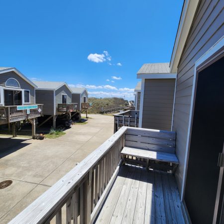 View from a wooden porch overlooking several gray cabins along a paved path under a sunny blue sky.