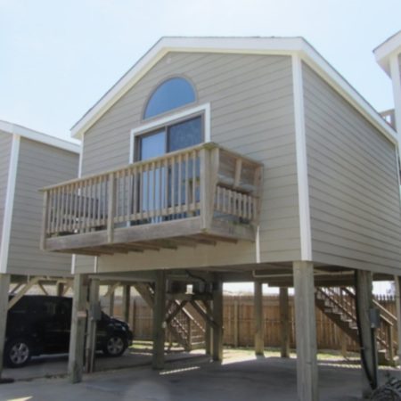 Elevated beach house with a wooden balcony, parked car underneath, and stairs leading up to the entrance.