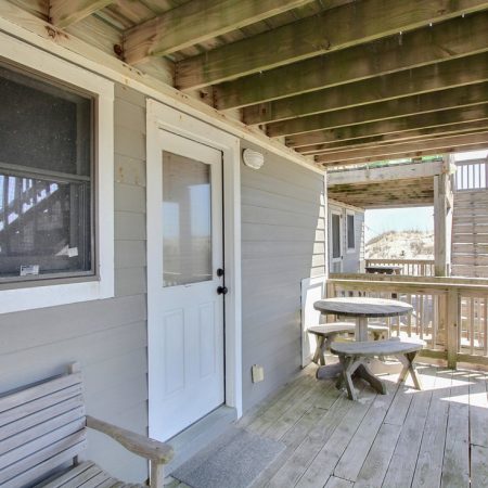 Wooden deck with a bench, round picnic tables, white door, window, and stairs leading to an upper level.