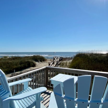 Two light blue Adirondack chairs on a deck overlook a sandy path leading to the beach and ocean under a clear sky.