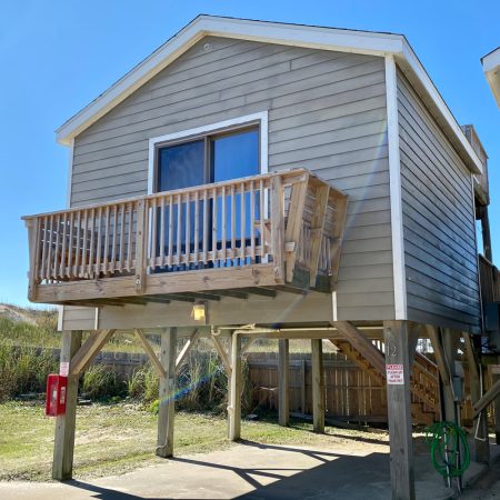 Elevated beige beach house with a small wooden balcony and exterior stairs, set on stilts above a concrete driveway.