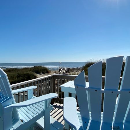 Two light blue Adirondack chairs on a wooden deck overlook the beach and ocean under a clear blue sky.