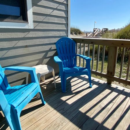 Two blue plastic chairs and a small table on a wooden deck, with sunlight casting shadows and greenery outside the railing.