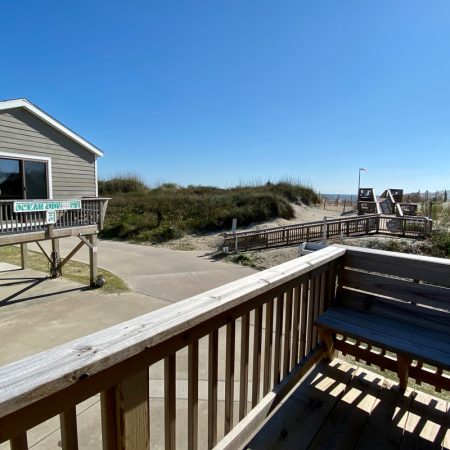 View from a porch overlooking a sandy path, grassy dunes, and a house with an “Ocean Dunes” sign under clear blue sky.