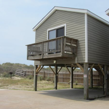Elevated gray beach house with a wooden balcony, supported by stilts, next to a sandy area and wooden fence.