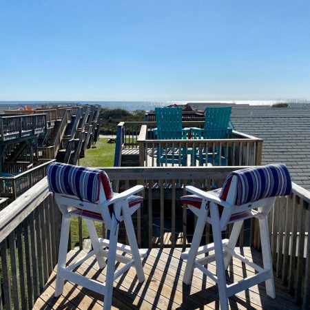 Two striped chairs on a wooden deck overlook rooftops, grassy yards, and the ocean under a clear blue sky.