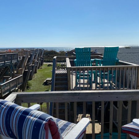 Wooden deck with striped cushioned chairs overlooks rooftops, elevated platforms, and the ocean in the background.