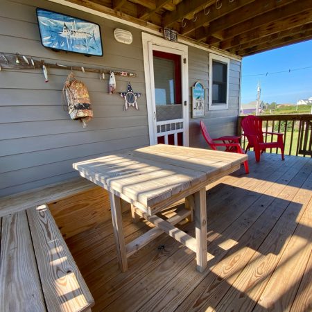 Wooden porch with a table, bench, two red chairs, and beach-themed wall decor in bright daylight.