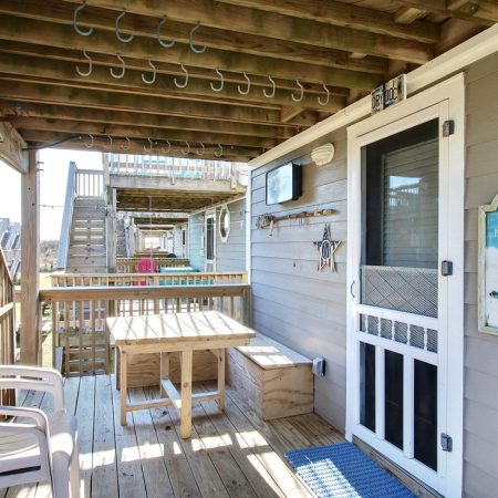 Second-story wooden deck with chairs, table, hooks on the ceiling, and a sign reading “Home is where the beach is.”.