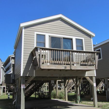 Gray elevated beach house on wooden stilts with a small balcony, stairs, and a parking area below.