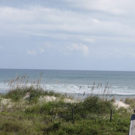 View of a sandy beach with sea grass, wooden railings, and ocean waves under a partly cloudy sky.