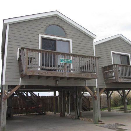 Gray stilt beach house with a wooden balcony and a sign reading “Cabana.” Driveway and stairs visible below.