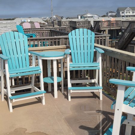 Two turquoise and white Adirondack chairs sit next to a round table on a wooden balcony overlooking houses.