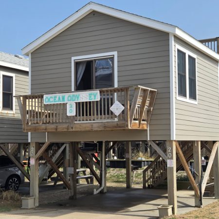 Gray beach house on stilts with a small balcony and a sign reading “Ocean Odyssey” above the entrance.