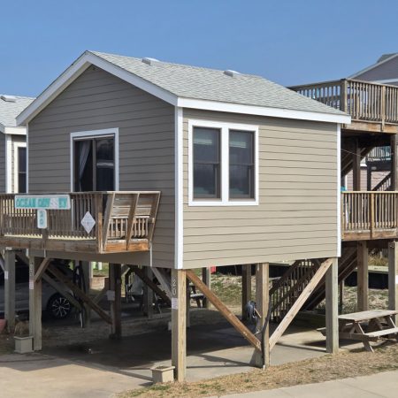 Beige stilt beach house with two decks, a staircase, and a picnic table beneath, next to similar elevated homes.