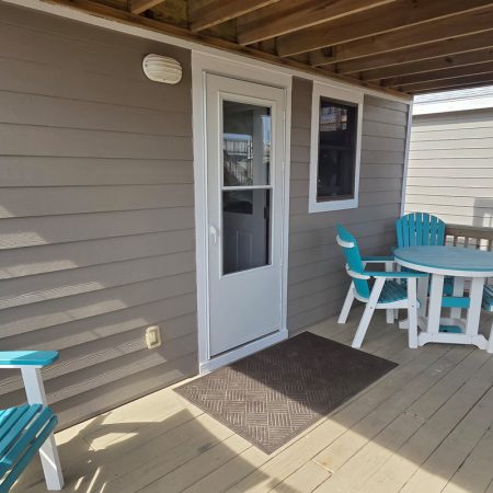 A covered porch with a white door, two turquoise chairs, and a round table on a wooden floor.