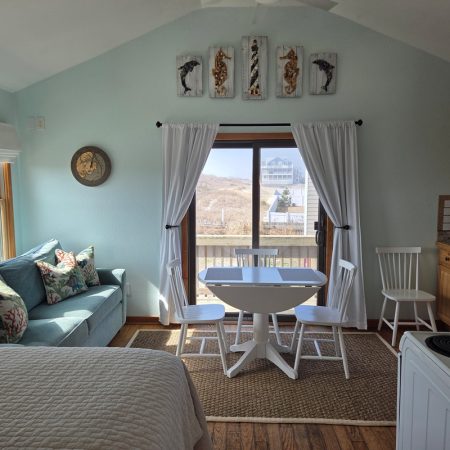 Sunny room with a sofa, table and chairs, bed, kitchen area, and large window overlooking a hilly landscape.