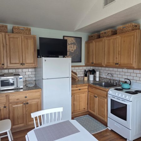 Small kitchen with wooden cabinets, white appliances, a microwave, fridge, stove, sink, table, and baskets on top shelves.