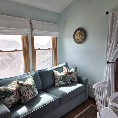Light blue sofa with patterned pillows by two windows; round wall clock above, dining table and chair nearby.