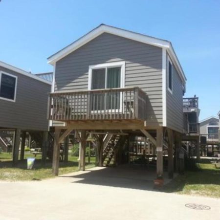 Gray stilt house with balcony and stairs, elevated above driveway, in a row of similar houses on a sunny day.