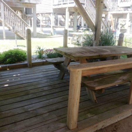Wooden picnic table with benches on a wooden deck under a raised structure, surrounded by greenery and stairs.