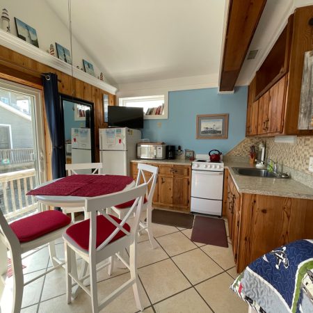 Small kitchen with wooden cabinets, white appliances, a dining table with red chairs, and a sliding glass door to a deck.