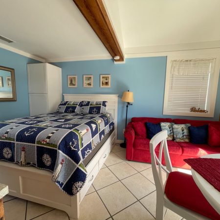 A small room with a bed, red sofa, white chairs, blue walls, and framed pictures, lit by sunlight.