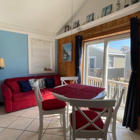A small sunlit dining area with a red couch, round table, and sliding glass door leading to an outdoor deck.