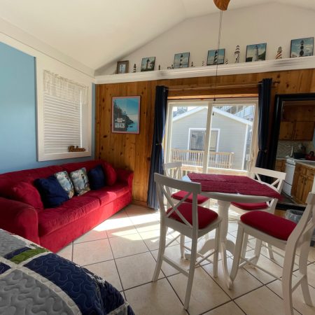 Small living area with a red couch, dining table, and kitchen; sunlight streams through glass doors to a patio.