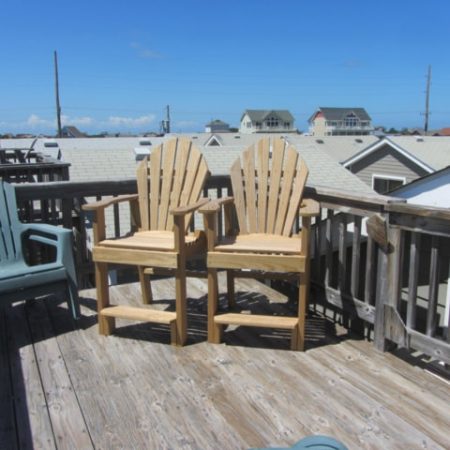 Three Adirondack chairs on a wooden deck with houses, blue sky, and rooftops in the background.