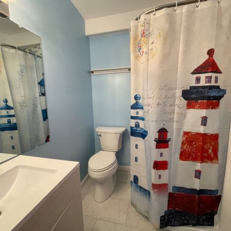Small bathroom with blue walls, white sink, and a lighthouse-themed shower curtain next to a toilet.