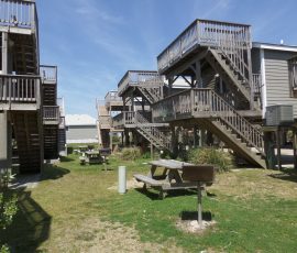 Wooden multi-level decks and staircases with picnic tables and grills on a grassy area under a clear sky.