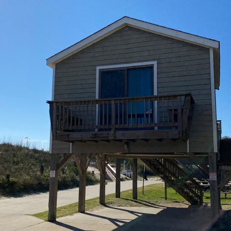 Elevated beach house with a small wooden balcony, stairs, and a sliding glass door, under a clear blue sky.