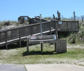 Outdoor wooden foot wash station near a boardwalk, surrounded by grass and sand dune vegetation under a clear sky.